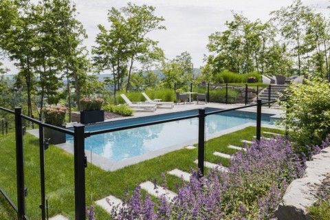 Two white long chairs on edge of in-ground swimming pool enclosed by clear glass and black metal