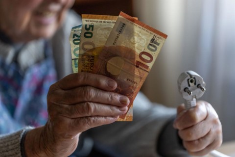 Senior citizen holding money and a power cable with plug in her hand at home, symbolising energy