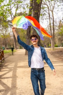 Vertical photo of a gay man raising and waving a rainbow colored hand fan in a park