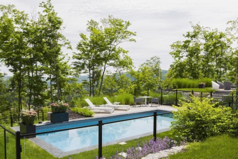 Two white long chairs on edge of in-ground swimming pool enclosed by clear glass and black metal