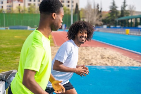 Side view of two smiling african young friends stretching and warming up in a running track