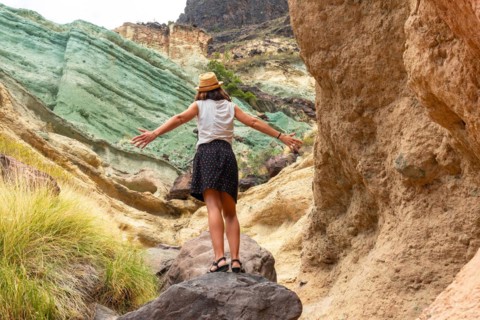 A tourist woman with a hat and open arms at the Azulejos de Veneguera or Rainbow Rocks in Mogan,