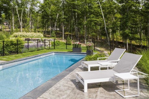 Two white long chairs on edge of in-ground swimming pool enclosed by clear glass and black metal