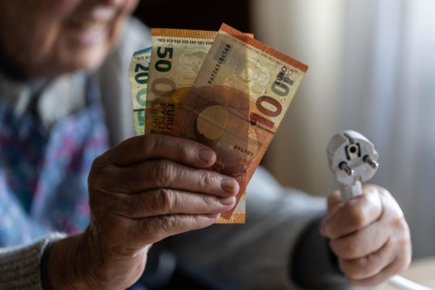 Senior citizen holding money and a power cable with plug in her hand at home, symbolising energy