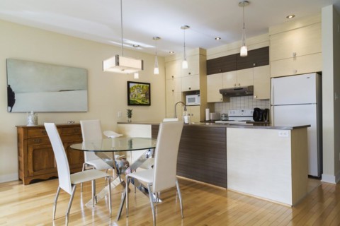Dining room with round glass table, white leather high -back chairs and kitchen inside a renovated