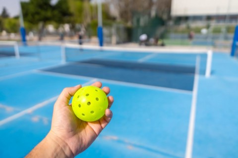 Unrecognizable person holding a plastic yellow pickleball ball with blue outdoor court on the