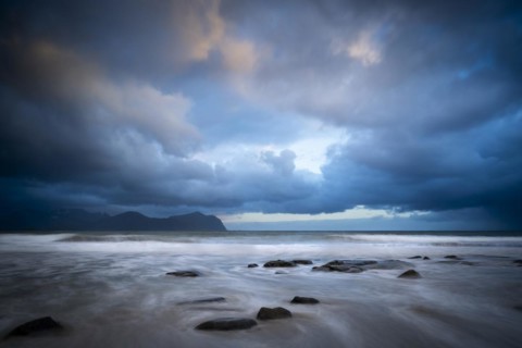 On the beach at Vikten. The sea washes around some rocks. Mountains in the background. At night at