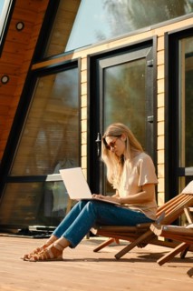 Woman checking mail on laptop sitting in a folding wooden chair on the terrace of summer house