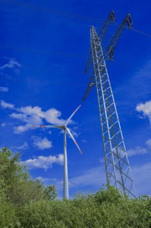 Power pylon with high-voltage lines and wind turbine behind green trees at the Avacon substation