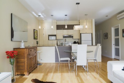 Dining room with round glass table, white leather high -back chairs and kitchen inside a renovated