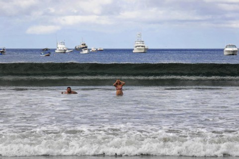 San Juan del Sur, Nicaragua, swimmers in the water in front of a big wave, boats and blue sky in