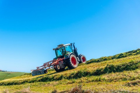A tractor is driving down a hillside, plowing the grass. The sky is clear and blue, and the sun is