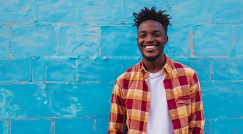 Black young positive man with dreadlocks is smiling and standing in front of the building wall.