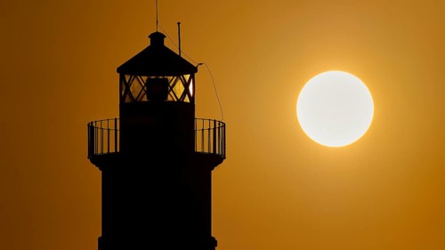 A lighthouse stands as a dark silhouette next to the round sun at sunrise, sunrise, dawn, Fort of