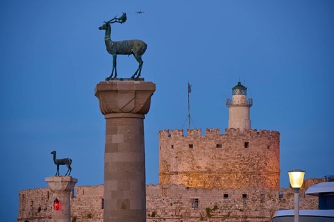Bronze deer on pedestal in the foreground with old fortress walls and lighthouse in the background,