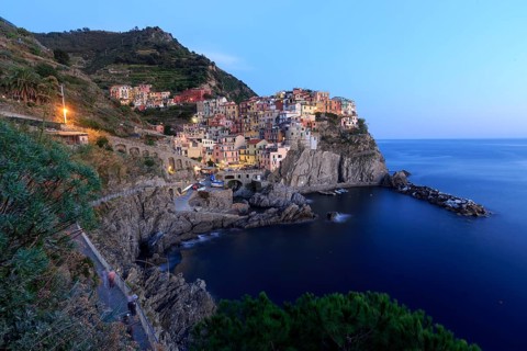 Mediterranean village on rocks on the coast at dusk with lights switched on and calm sea, Italy,