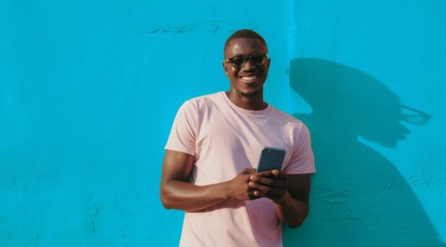 Black young positive man with dreadlocks is smiling and standing in front of the building wall.
