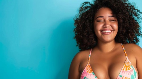 A plus size black afro-american bikini model with curly hair is smiling and wearing a bikini top.