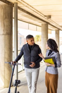 Woman in formal suit talking to man holding a scooter. Girl in formal suit talking to man holding