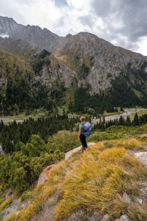 A female hiker stands on a grassy slope against an impressive mountain backdrop under a cloudy sky,