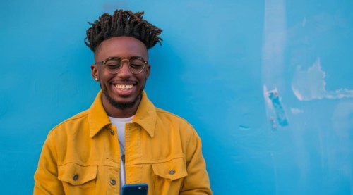 Black young positive man with dreadlocks is smiling and standing in front of the building wall.