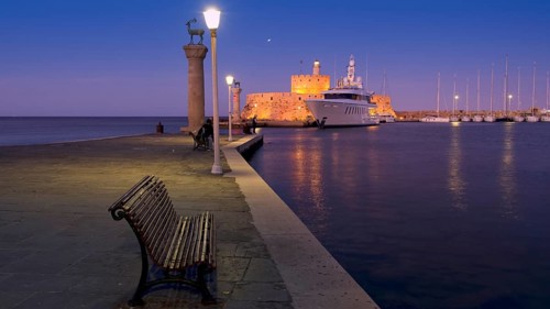 Quiet harbour promenade with bench and lamppost opposite fort and boats in moonlight, night shot,