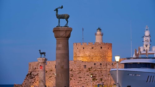 A deer sculpture on a pillar in front of a castle and lighthouse next to a luxury yacht at dusk,