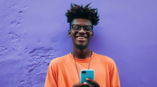 Black young positive man with dreadlocks is smiling and standing in front of the building wall.