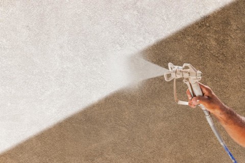 Professional spray painter holding spray gun spraying white paint over brown stucco surface