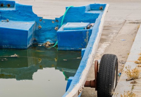 Small blue fishing boat missing engine on trailer full of water with a green hose at aft to drain