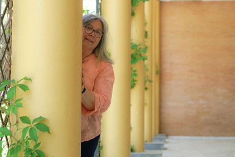 Older woman with white hair and glasses, smiling from behind a column