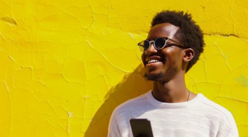 Black young positive man with dreadlocks is smiling and standing in front of the building wall.