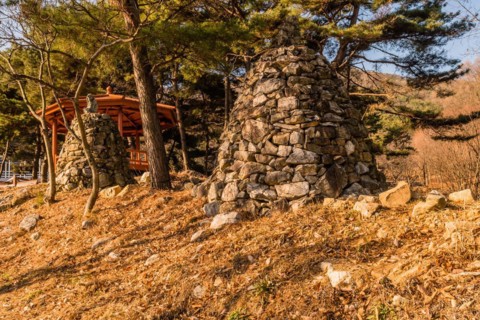 Two stone pyramids on small hill under grove of evergreen trees and wooden gazebo in background