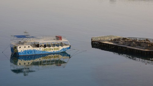 An excursion boat is reflected in the calm waters next to a wooden jetty, Lindos, Rhodes,