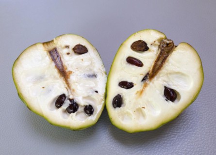 A sliced Chirimuya with visible dark seeds lies on a grey surface, chalklike texture on a light