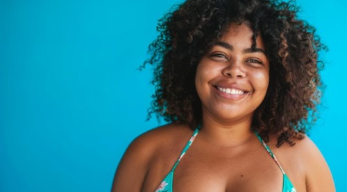 A plus size black afro-american bikini model with curly hair is smiling and wearing a bikini top.