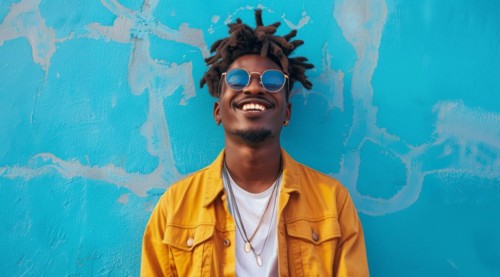 Black young positive man with dreadlocks is smiling and standing in front of the building wall.