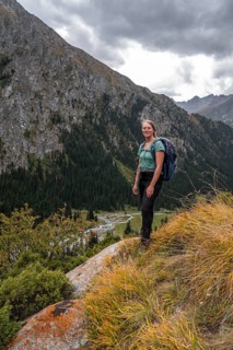 A female hiker stands on a grassy slope against an impressive mountain backdrop under a cloudy sky,