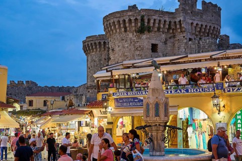 Hippocrates Square, St Paul's Gate, visitors walk between restaurants and shops by an old castle,