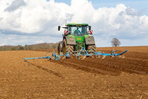 Green John Deere tractor ploughing deep furrows to prepare soil for potato crop, Ramsholt, Suffolk,