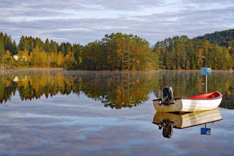 Trees and a small boat reflected in the calm waters of a lake, leaf colouring, Bullaren, Bohuslaen.