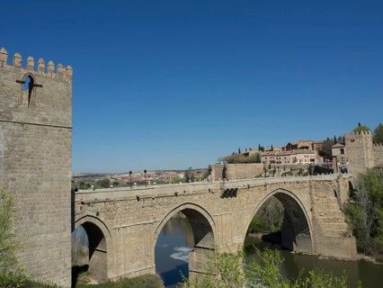 Historic stone bridge over a river with city walls and buildings in the background under a sunny