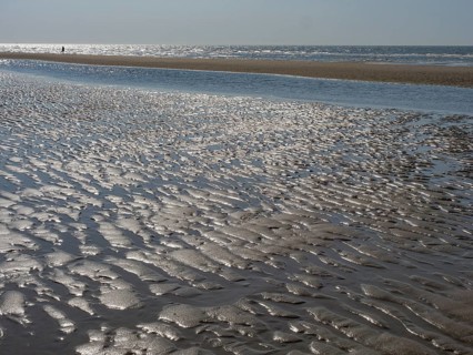 Close-up of a sandy beach with watercourse and waves in the background, glittering sea water at low