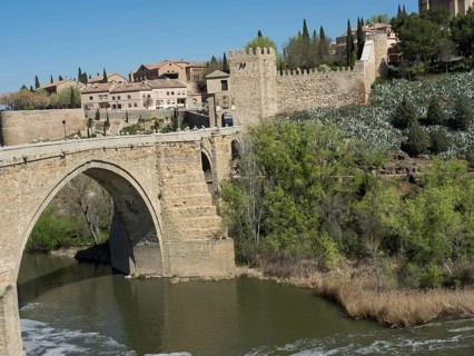 Stone bridge over a river in front of a castle wall and a city view with vegetation, blue sky,