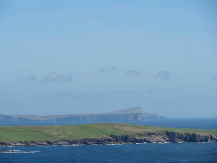 Wide green fields meet the deep blue sea, with a visible island in the background, green meadows on