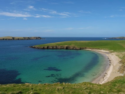 Small bay with clear water and a sandy beach surrounded by green grassland and a wide blue sky,