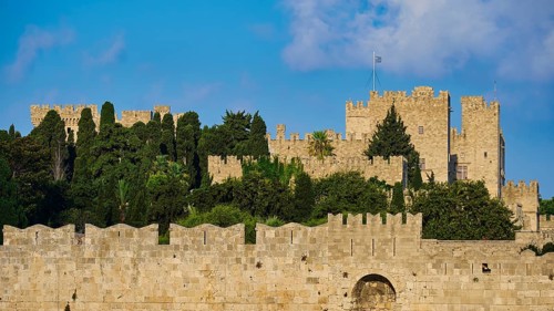 Fortress walls with battlements under a bright blue sky, surrounded by green vegetation, part of