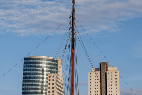 Sail mast between two modern buildings in front of a blue sky, skyline of a modern city on a river