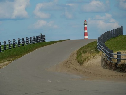 Road leads slightly uphill to a lighthouse, flanked by fences, with cloudy sky, red and white