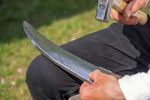 Man peening a scythe blade using a jig and a hammer to sharpen it